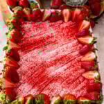 Overhead view of a strawberry tres leches cake topped with whipped cream and a vibrant layer of strawberry powder, decorated with fresh halved strawberries around the edges in a rectangular baking dish.