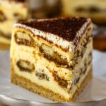 A close-up slice of tiramisu cheesecake on a white plate, showing layers of espresso-soaked ladyfingers, creamy cheesecake filling, and a graham cracker crust, all topped with a dusting of cocoa powder.