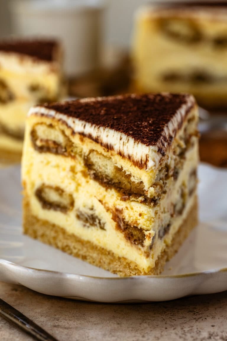 A close-up slice of tiramisu cheesecake on a white plate, showing layers of espresso-soaked ladyfingers, creamy cheesecake filling, and a graham cracker crust, all topped with a dusting of cocoa powder.