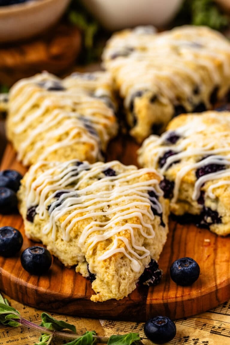 A close-up of golden blueberry scones drizzled with sweet glaze, arranged on a wooden board with fresh blueberries scattered around. The scones have a tender, crumbly texture with juicy blueberries peeking through, and the scene includes rustic sheet music and fresh greenery in the background.