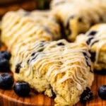 A close-up of golden blueberry scones drizzled with sweet glaze, arranged on a wooden board with fresh blueberries scattered around. The scones have a tender, crumbly texture with juicy blueberries peeking through, and the scene includes rustic sheet music and fresh greenery in the background.