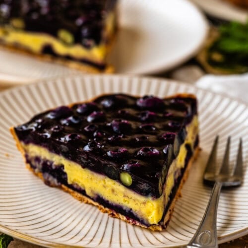 A close-up slice of blueberry tart on a striped plate, featuring a golden crust, creamy vanilla custard, and a glossy blueberry topping, with a fork and fresh mint beside the plate.