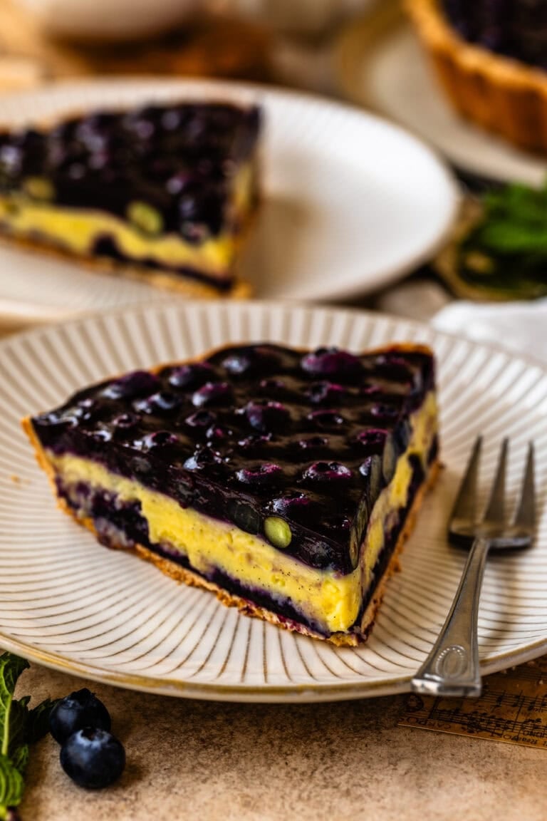 A close-up slice of blueberry tart on a striped plate, featuring a golden crust, creamy vanilla custard, and a glossy blueberry topping, with a fork and fresh mint beside the plate.