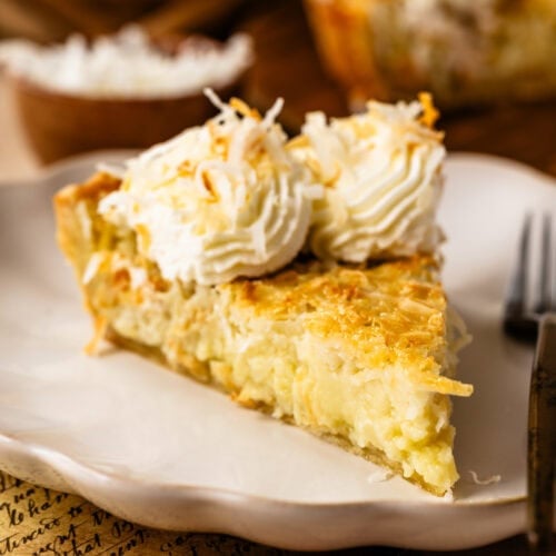 A close-up of a slice of coconut cream pie on a white plate, with a golden, flaky crust, creamy coconut filling, and piped whipped cream topped with toasted coconut.