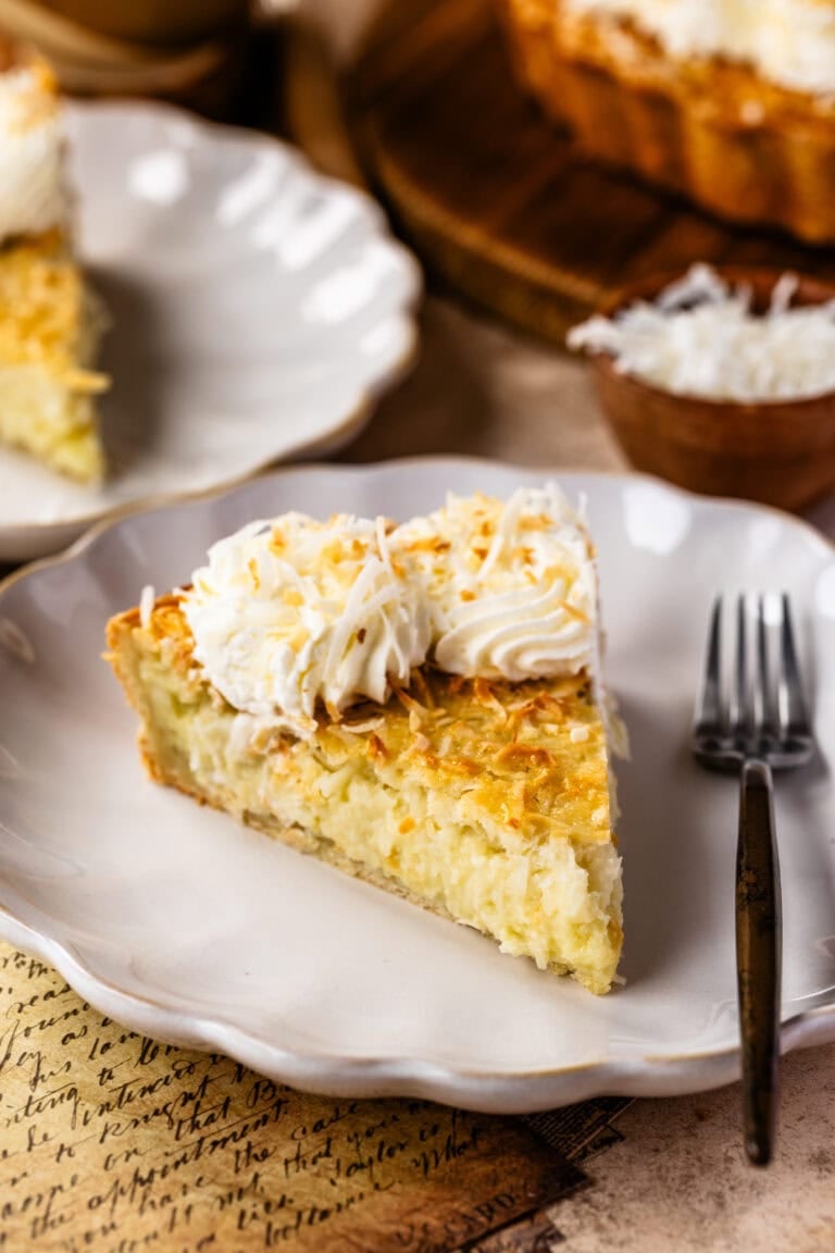 A close-up of a slice of coconut cream pie on a white plate, with a golden, flaky crust, creamy coconut filling, and piped whipped cream topped with toasted coconut.