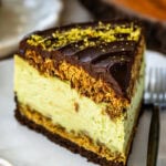 A close-up of a single slice of Dubai Chocolate Cheesecake on a white plate, showcasing its layers: a chocolate cookie crust, creamy pistachio cheesecake filling, toasted kataifi, and glossy chocolate ganache topped with crushed pistachios. A fork rests beside the slice, with a soft-focus background featuring rustic decor and another slice of cheesecake.