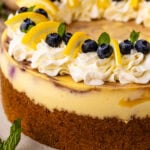 Close-up of a lemon blueberry cheesecake topped with whipped cream, fresh blueberries, lemon slices, and mint leaves, with a graham cracker crust base.