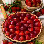 A whole strawberry tart with a golden crust and vibrant glazed strawberries on top, displayed on a wooden board with fresh strawberries and rustic decor in the background.