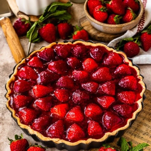A whole strawberry tart with a golden crust and vibrant glazed strawberries on top, displayed on a wooden board with fresh strawberries and rustic decor in the background.