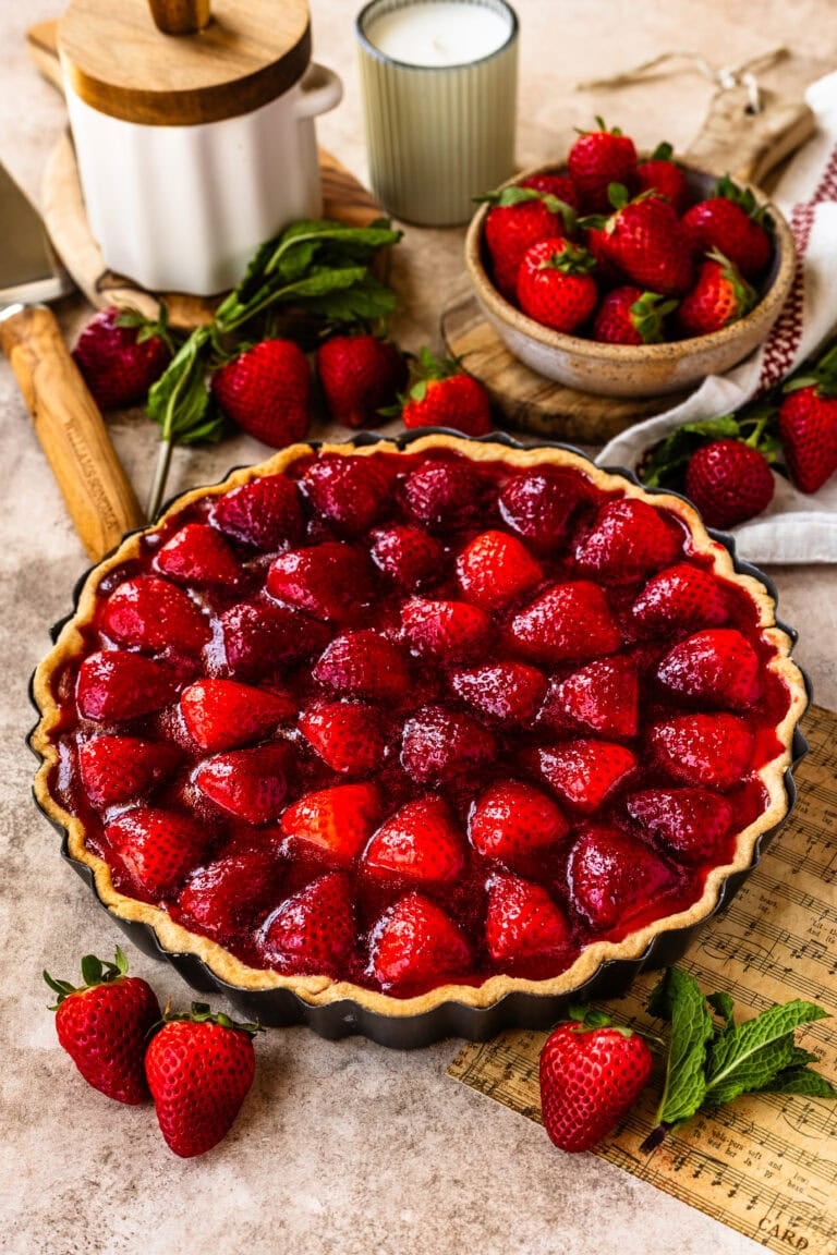 A whole strawberry tart with a golden crust and vibrant glazed strawberries on top, displayed on a wooden board with fresh strawberries and rustic decor in the background.