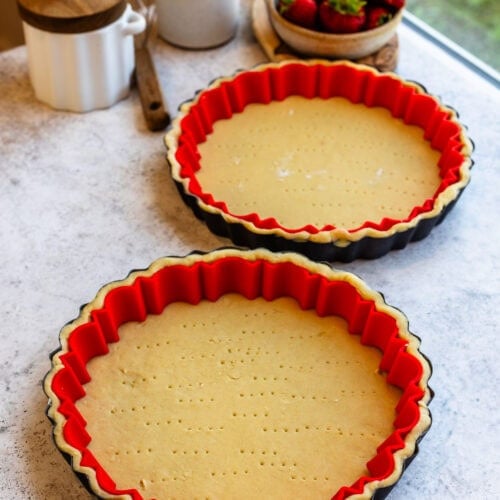 Two unbaked tart crusts in fluted tart pans lined with red silicone edge protectors, resting on a light countertop with fresh strawberries and rustic decor in the background.