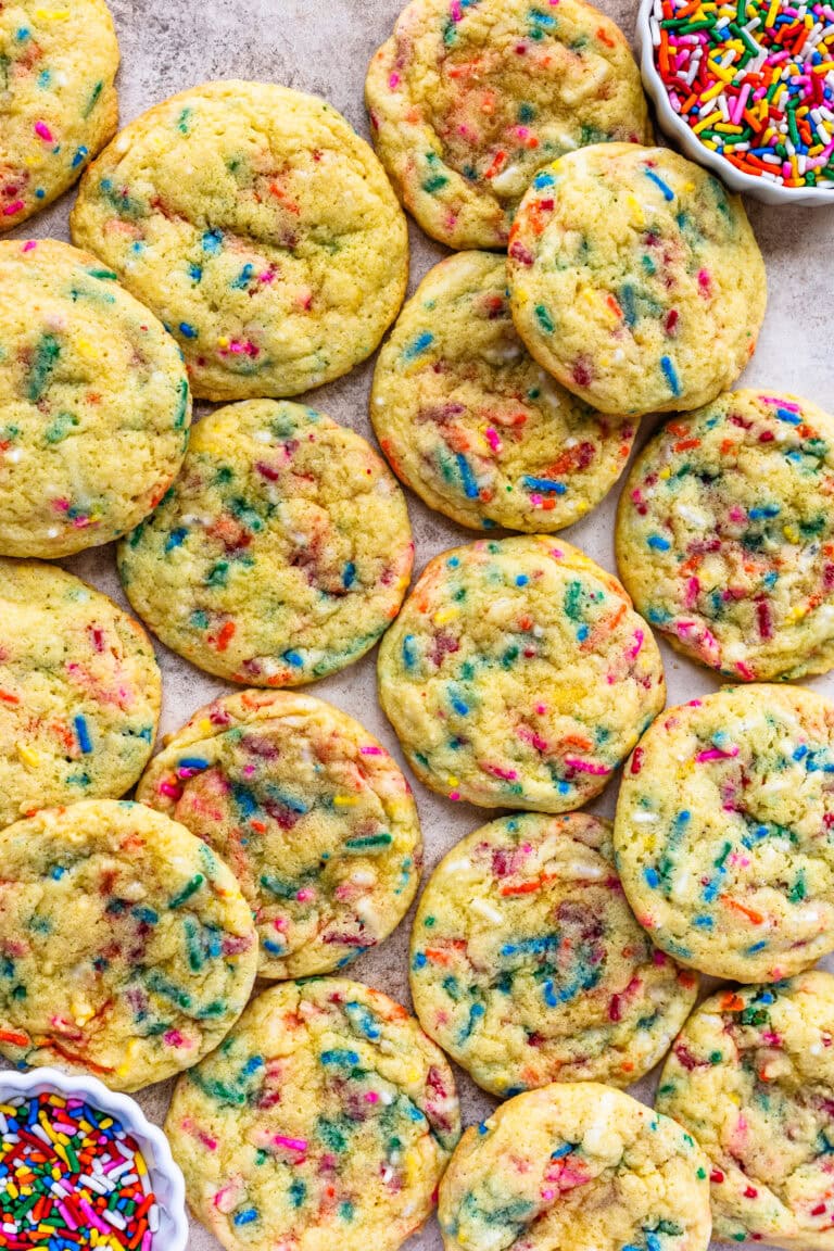 A batch of vibrant Funfetti cookies spread out on a surface, showcasing colorful sprinkles throughout, with a small bowl of rainbow sprinkles on the side.