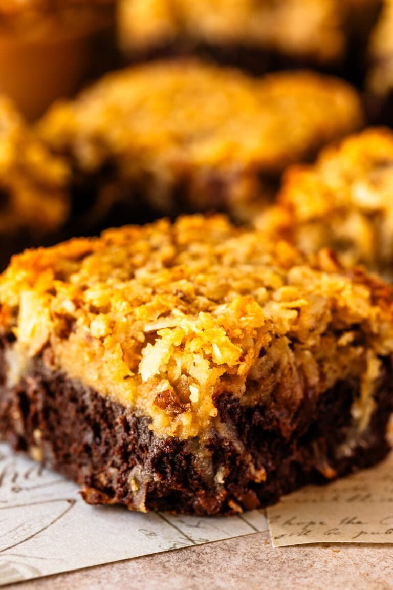 A close-up of a German chocolate brownie showing its rich, fudgy base and thick, golden coconut-pecan topping with crispy toasted edges.