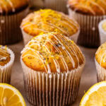A close-up of golden lemon poppyseed muffins topped with a drizzle of icing and fresh lemon zest, surrounded by halved lemons on a neutral background.