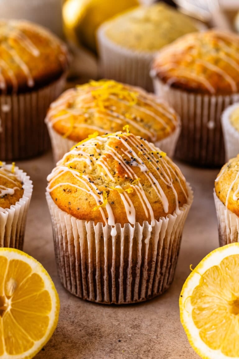 A close-up of golden lemon poppyseed muffins topped with a drizzle of icing and fresh lemon zest, surrounded by halved lemons on a neutral background.