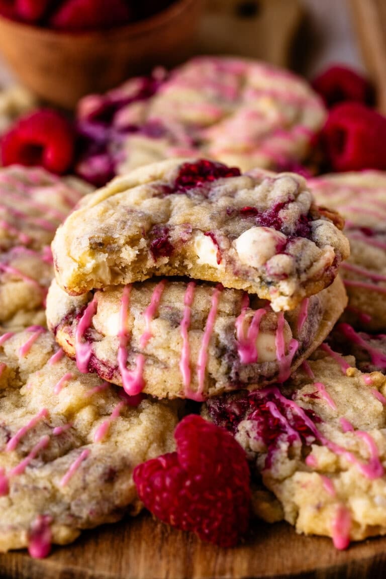 Close-up of raspberry white chocolate cookies stacked on a wooden board, with the top cookie showing a gooey bite taken out. The cookies are drizzled with pink icing and surrounded by fresh raspberries.