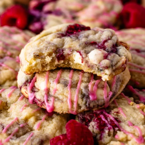 Close-up of raspberry white chocolate cookies drizzled with pink icing and surrounded by fresh raspberries on a wooden board. The cookies have golden edges and vibrant berry swirls.