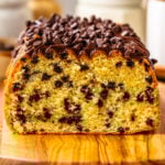 A close-up of a chocolate chip loaf cake topped with chocolate ganache and mini chips, sliced to reveal its soft, golden crumb and rich chocolate studding.