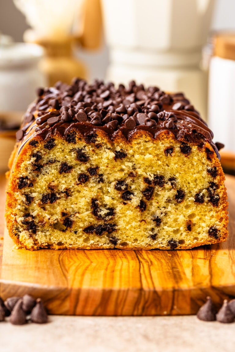 A close-up of a chocolate chip loaf cake topped with chocolate ganache and mini chips, sliced to reveal its soft, golden crumb and rich chocolate studding.