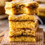 A stack of three golden cinnamon roll blondies with a visible cinnamon-sugar swirl in the center and a drizzle of glaze on top. One has a bite taken out of it. The bars are arranged on a round wooden board, with more blondies in the background and cinnamon sticks off to the side.