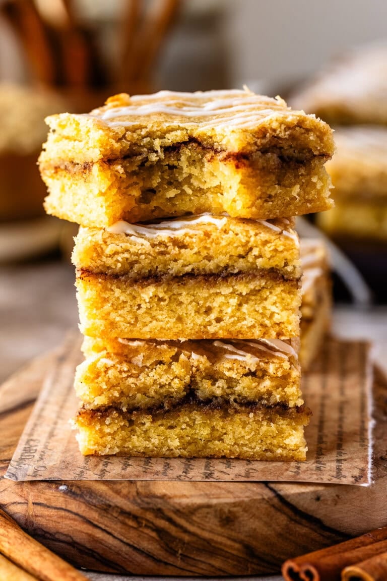 A stack of three golden cinnamon roll blondies with a visible cinnamon-sugar swirl in the center and a drizzle of glaze on top. One has a bite taken out of it. The bars are arranged on a round wooden board, with more blondies in the background and cinnamon sticks off to the side.