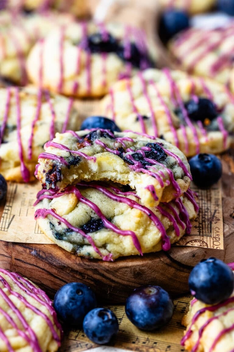 Soft blueberry cookies drizzled with vibrant purple glaze, surrounded by fresh blueberries on a piece of decorative parchment paper.