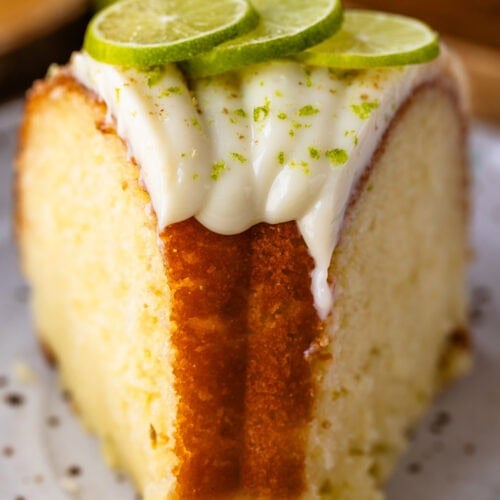 A close-up of a slice of lime bundt cake topped with piped lime glaze, fresh lime slices, and a sprinkle of lime zest, showing the cake’s soft, moist crumb and golden edges.