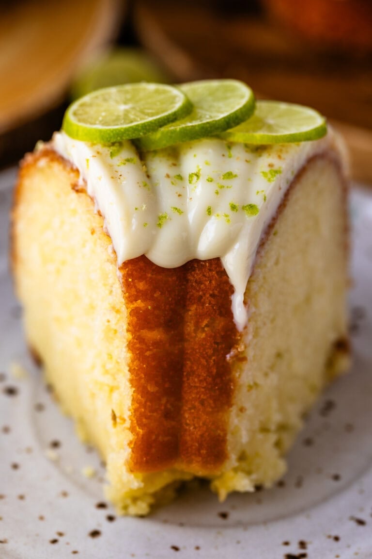 A close-up of a slice of lime bundt cake topped with piped lime glaze, fresh lime slices, and a sprinkle of lime zest, showing the cake’s soft, moist crumb and golden edges.