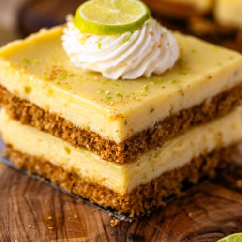 A close-up of two stacked key lime pie bars on a wooden board, each with a thick graham cracker crust, creamy lime filling, and a swirl of whipped cream topped with a fresh lime slice.