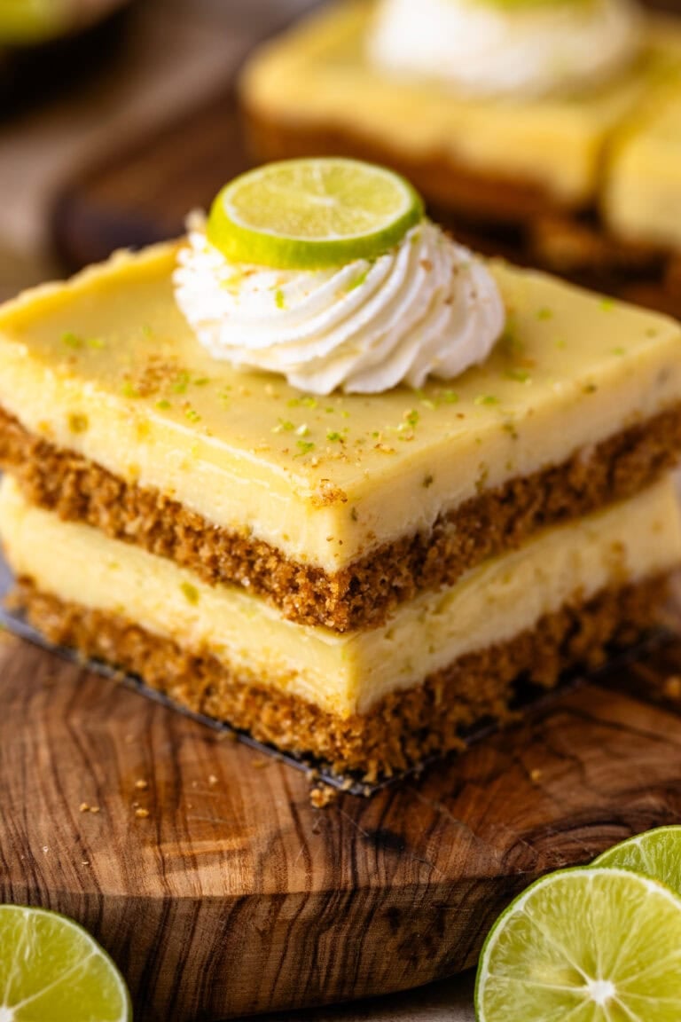A close-up of two stacked key lime pie bars on a wooden board, each with a thick graham cracker crust, creamy lime filling, and a swirl of whipped cream topped with a fresh lime slice.