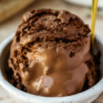A dish of creamy homemade chocolate ice cream, scooped high in a glass dessert bowl with a spoon, slightly melting for the perfect just-scooped look.