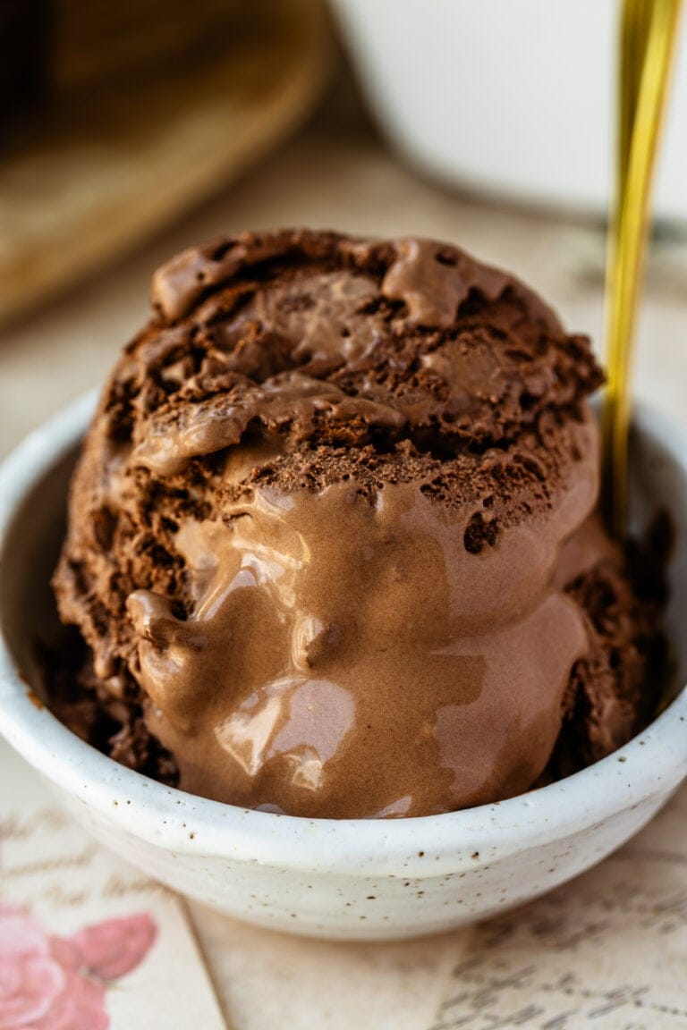 A dish of creamy homemade chocolate ice cream, scooped high in a glass dessert bowl with a spoon, slightly melting for the perfect just-scooped look.