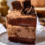 A tall slice of chocolate ice cream cake with layers of chocolate cake, chocolate ice cream, and chocolate frosting, topped with a brownie square and served on a speckled ceramic plate.