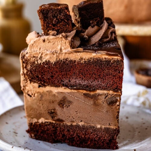 A tall slice of chocolate ice cream cake with layers of chocolate cake, chocolate ice cream, and chocolate frosting, topped with a brownie square and served on a speckled ceramic plate.