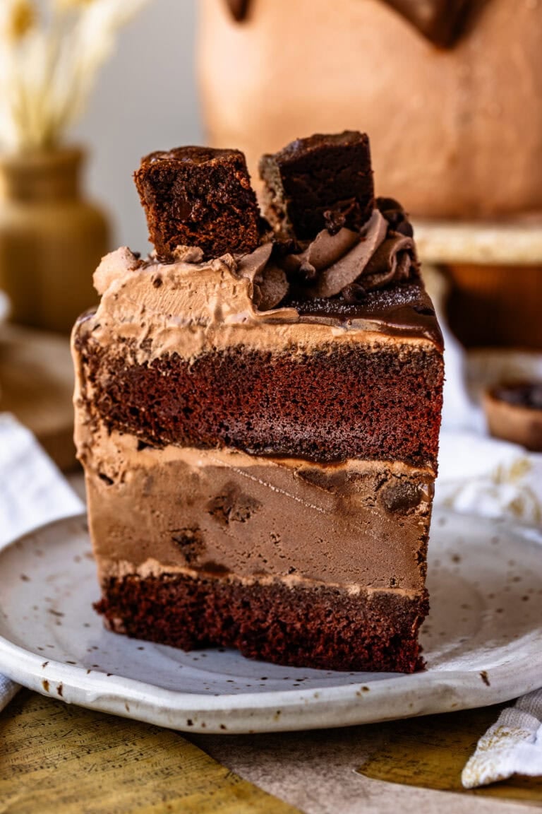 A tall slice of chocolate ice cream cake with layers of chocolate cake, chocolate ice cream, and chocolate frosting, topped with a brownie square and served on a speckled ceramic plate.