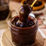 A small glass jar filled with thick, glossy hot fudge sauce and a gold spoon dipped inside, with a second jar blurred in the background.
