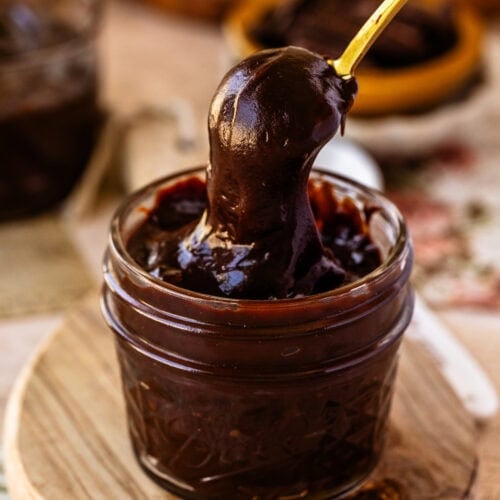 A small glass jar filled with thick, glossy hot fudge sauce and a gold spoon dipped inside, with a second jar blurred in the background.
