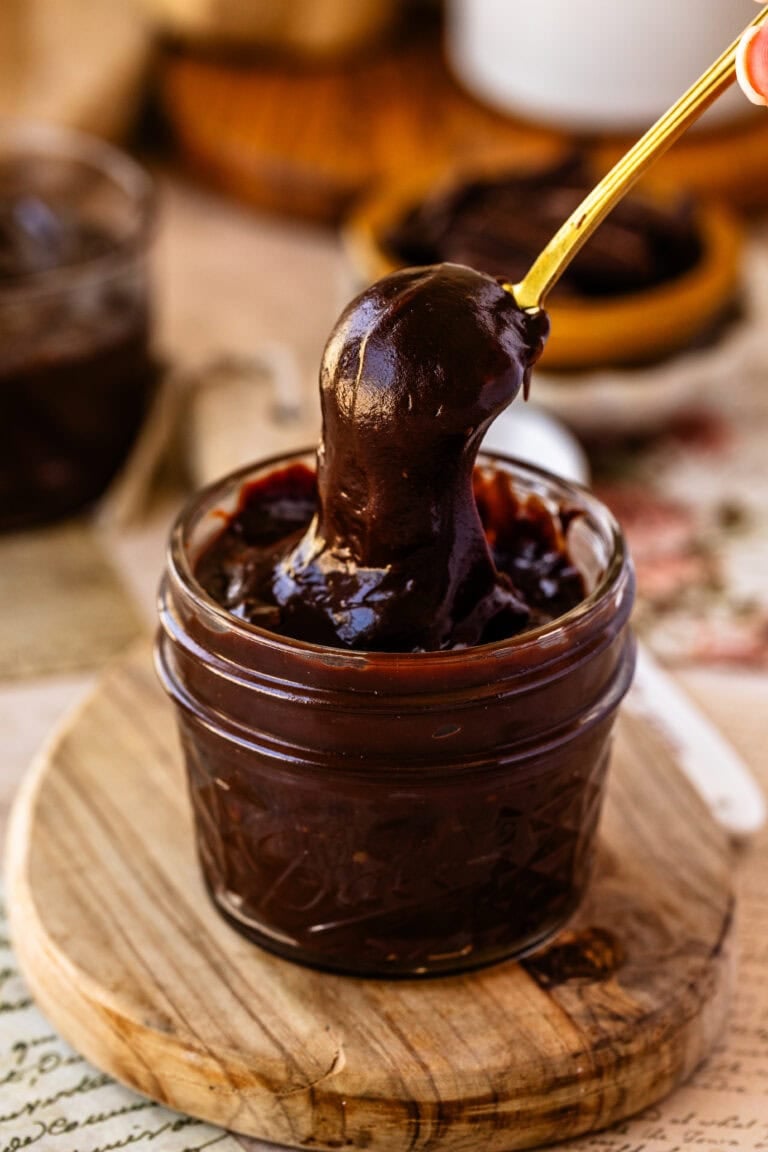 A small glass jar filled with thick, glossy hot fudge sauce and a gold spoon dipped inside, with a second jar blurred in the background.