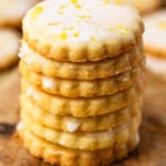 A stack of glazed lemon shortbread cookies on a wooden board, with flecks of lemon zest on top and crisp, golden edges.