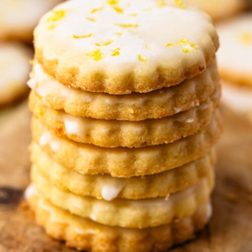A stack of glazed lemon shortbread cookies on a wooden board, with flecks of lemon zest on top and crisp, golden edges.