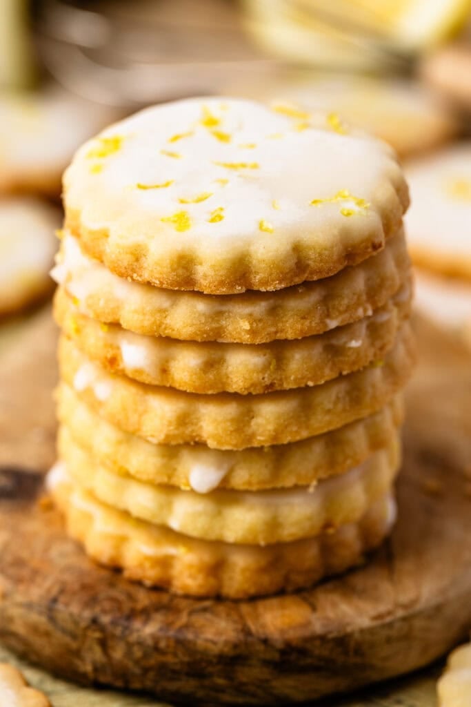 A stack of glazed lemon shortbread cookies on a wooden board, with flecks of lemon zest on top and crisp, golden edges.