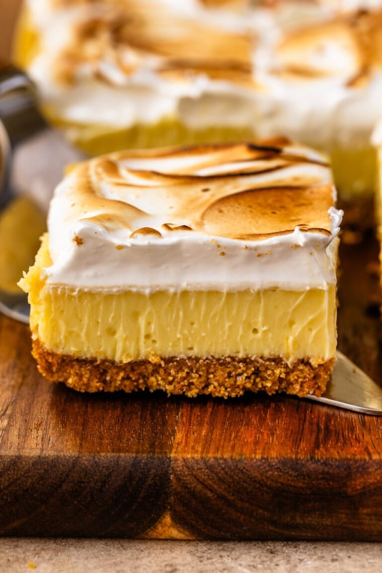 A close-up of a lemon meringue pie slice with a graham cracker crust, creamy lemon filling, and a thick layer of toasted meringue on top.