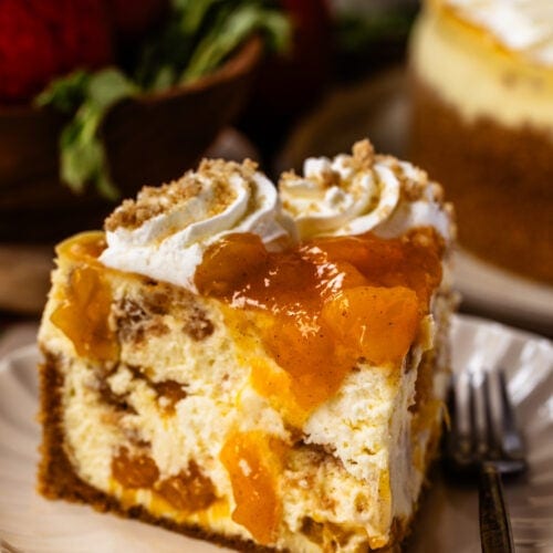 A close-up slice of peach cobbler cheesecake on a scalloped plate, topped with peach compote, whipped cream swirls, and crumbled streusel, with a fork resting beside it.