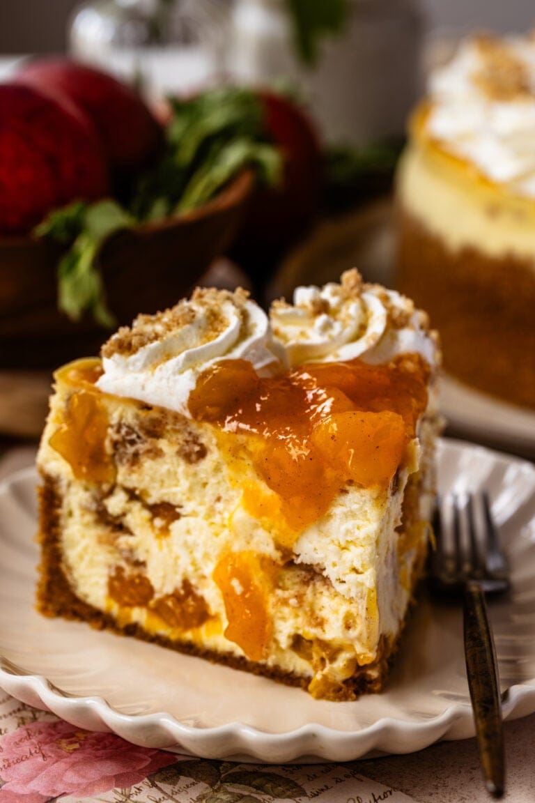A close-up slice of peach cobbler cheesecake on a scalloped plate, topped with peach compote, whipped cream swirls, and crumbled streusel, with a fork resting beside it.