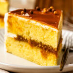 A close-up of a slice of vanilla caramel cake with layers of moist vanilla cake, rich caramel filling, vanilla buttercream, and a glossy caramel topping garnished with chocolate pearls.