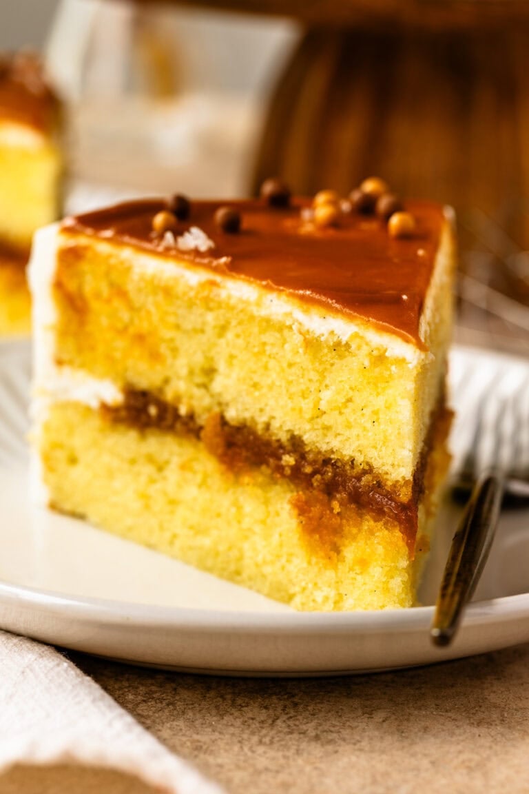A close-up of a slice of vanilla caramel cake with layers of moist vanilla cake, rich caramel filling, vanilla buttercream, and a glossy caramel topping garnished with chocolate pearls.