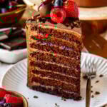 A slice of 8-layer chocolate cake on a white plate, showing thin chocolate layers filled with buttercream and berry jam, topped with fresh berries and chocolate shavings.