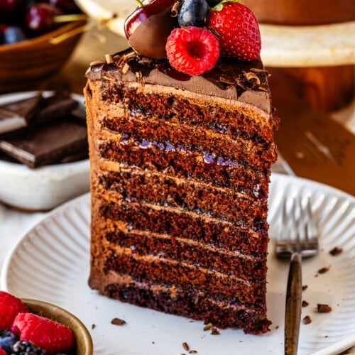 A slice of 8-layer chocolate cake on a white plate, showing thin chocolate layers filled with buttercream and berry jam, topped with fresh berries and chocolate shavings.