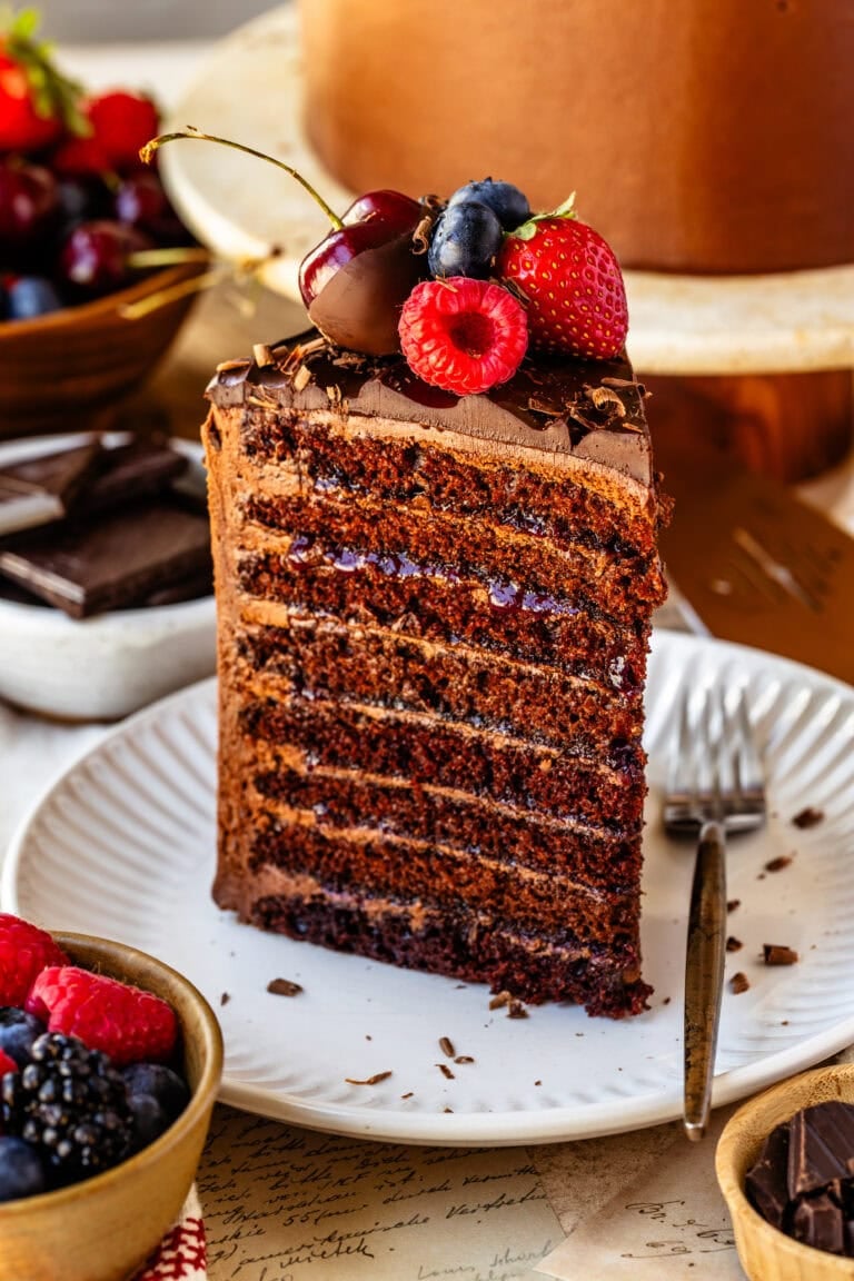 A slice of 8-layer chocolate cake on a white plate, showing thin chocolate layers filled with buttercream and berry jam, topped with fresh berries and chocolate shavings.