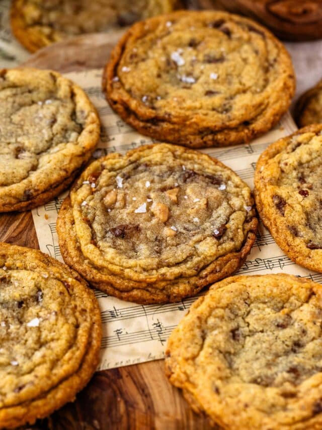 A close-up of freshly baked toffee brown butter cookies topped with flaky sea salt, arranged on a wooden surface with sheet music underneath.